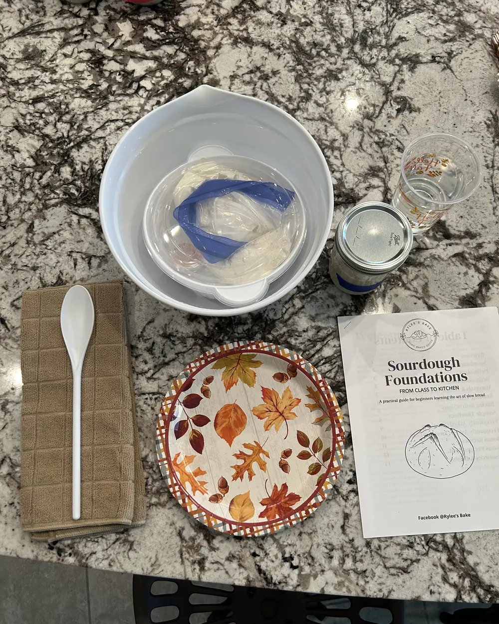 A class place setting: mixing bowl with starter, spoon, tea towel and the Sourdough Foundations guide