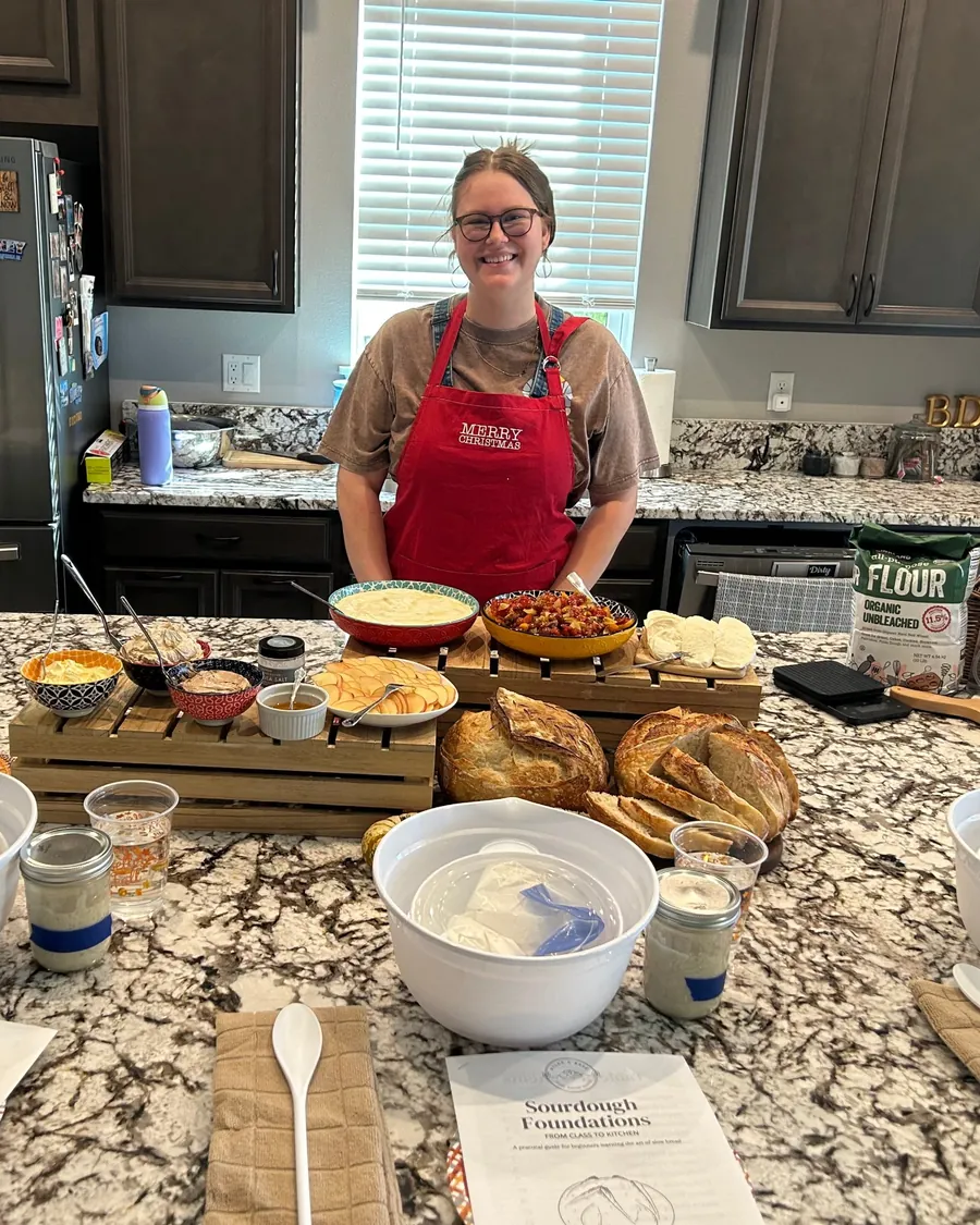 Rylee in a red apron, smiling behind a class table of fresh sourdough and appetizers